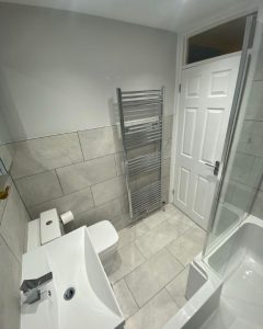 A new bathroom installation featuring grey tiled walls and floors, shower over bath and a new WC and sink.