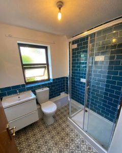 A new bathroom installation featuring vinyl flooring, blue tiled walls, a walk-in shower enclosure and a new WC and sink.