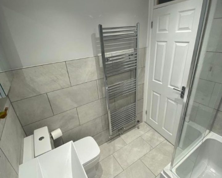 A new bathroom installation featuring grey tiled walls and floors, shower over bath and a new WC and sink.